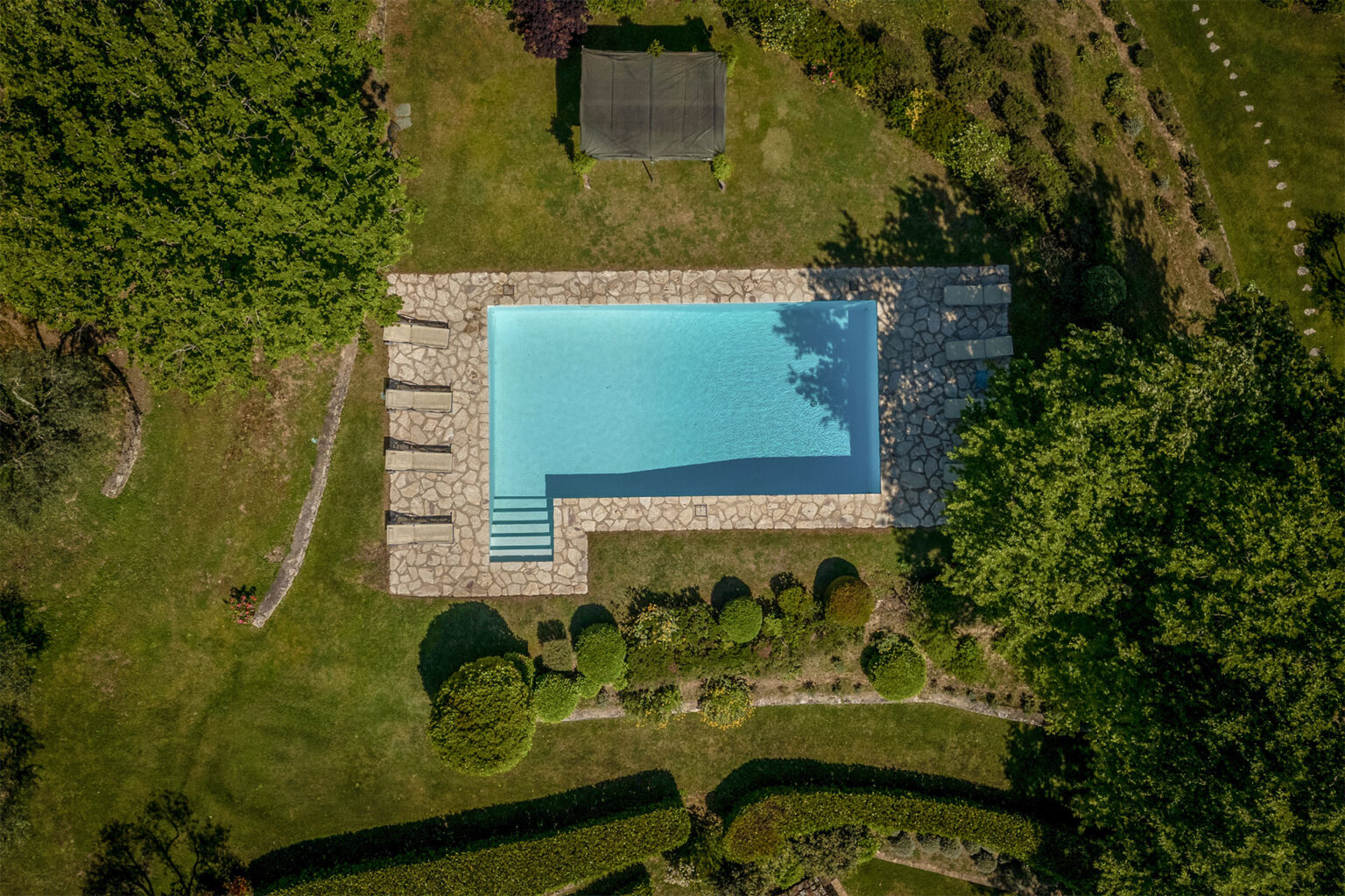 Aerial view of a serene backyard swimming pool surrounded by lush greenery and landscaped gardens, featuring stone decking and steps leading into the water. Ideal for relaxation and outdoor gatherings. | Only on SopranoVillas
