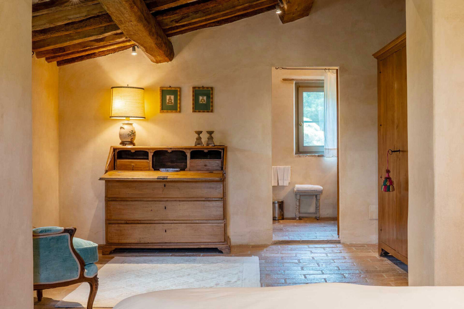 Cozy rustic bedroom featuring exposed wooden beams, a vintage wooden dresser, a decorative lamp, and framed artwork on the walls. A doorway leads to a bright bathroom with natural light, while a plush armchair adds a touch of comfort to the space. | Only on SopranoVillas