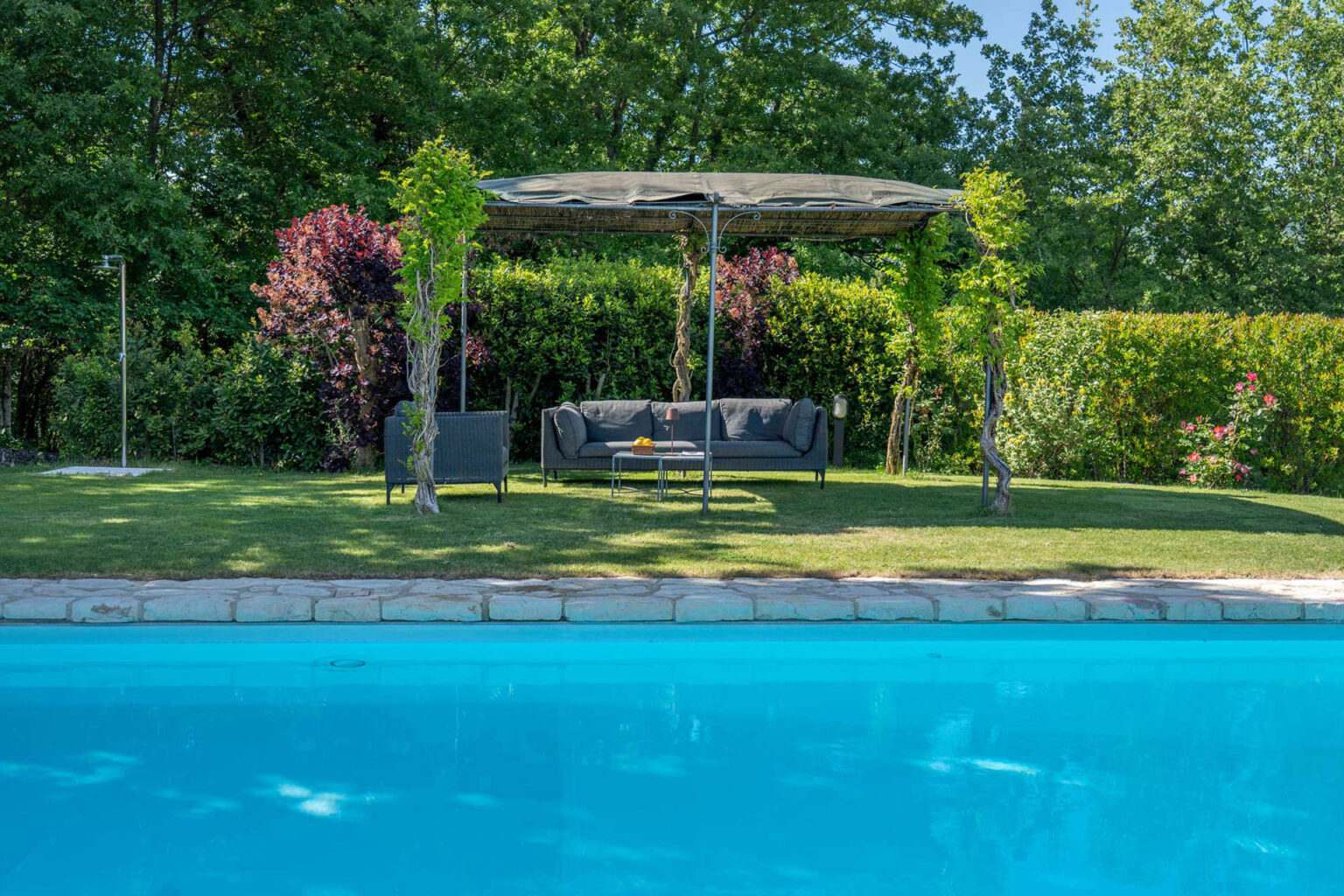 Relaxing outdoor lounge area by a swimming pool, featuring a shaded sofa set under a canopy, surrounded by lush greenery and colorful shrubs. | Only on SopranoVillas