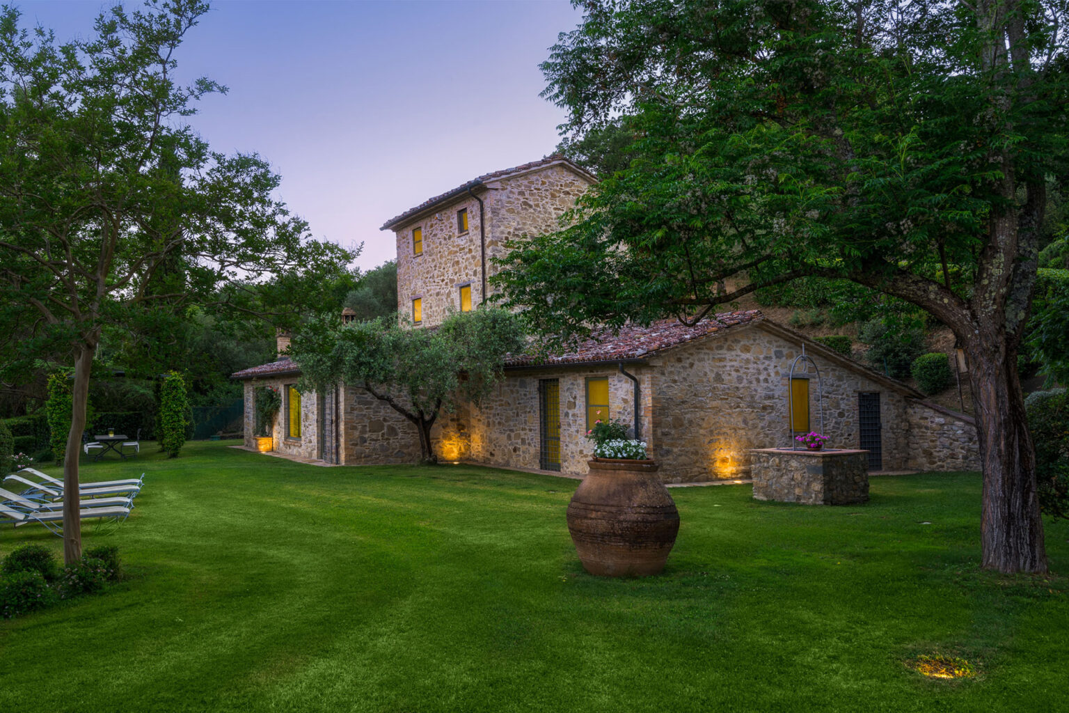 Charming stone villa surrounded by lush greenery at dusk, featuring a beautifully landscaped garden and warm exterior lighting. | Only on SopranoVillas