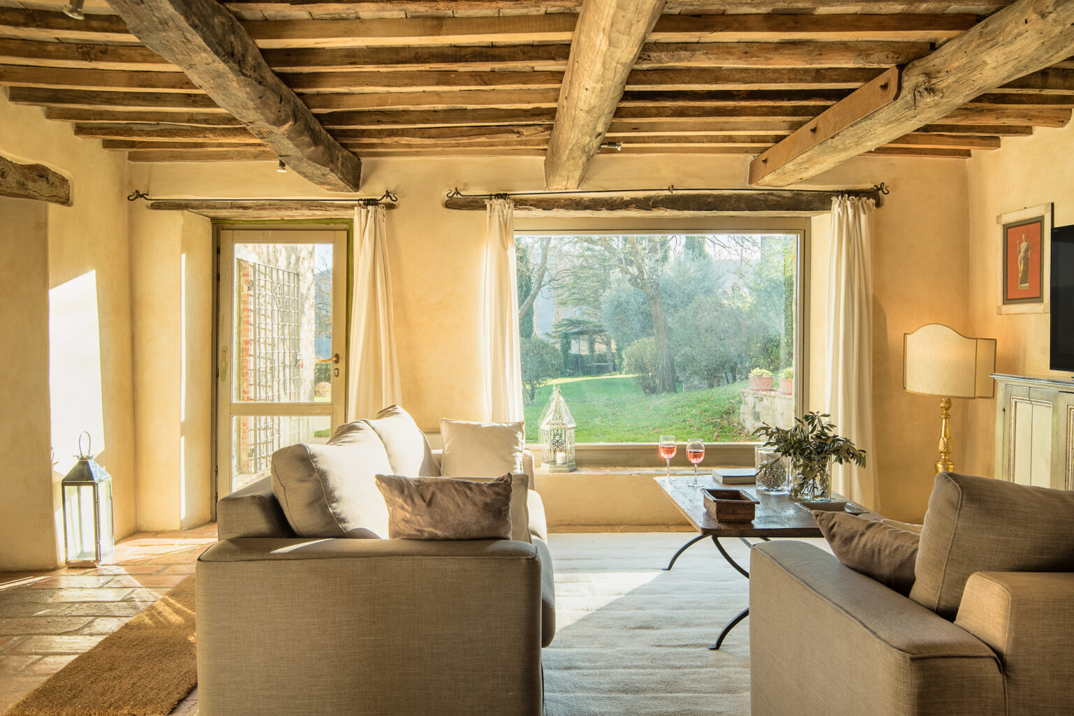 Cozy living room with exposed wooden beams, large windows offering a view of a garden, and comfortable gray sofas. Natural light floods the space, highlighting a stylish coffee table and decorative elements. | Only on SopranoVillas