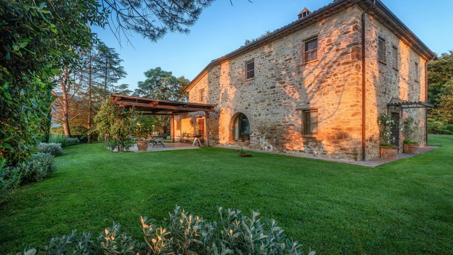 Historic stone villa surrounded by lush green lawn and trees, showcasing rustic architecture and a cozy outdoor patio area in the warm evening light. | Only on SopranoVillas