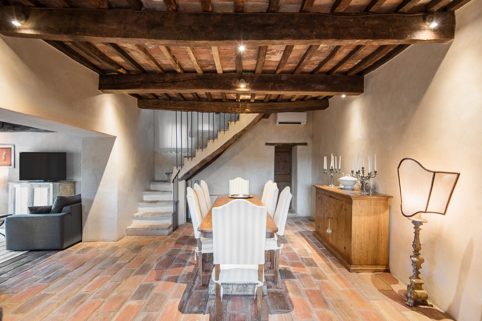 Spacious dining area with a wooden dining table and white chairs, featuring rustic beams and warm lighting, leading to a staircase. The decor includes a wooden sideboard and a stylish lamp, creating a cozy atmosphere. | Only on SopranoVillas