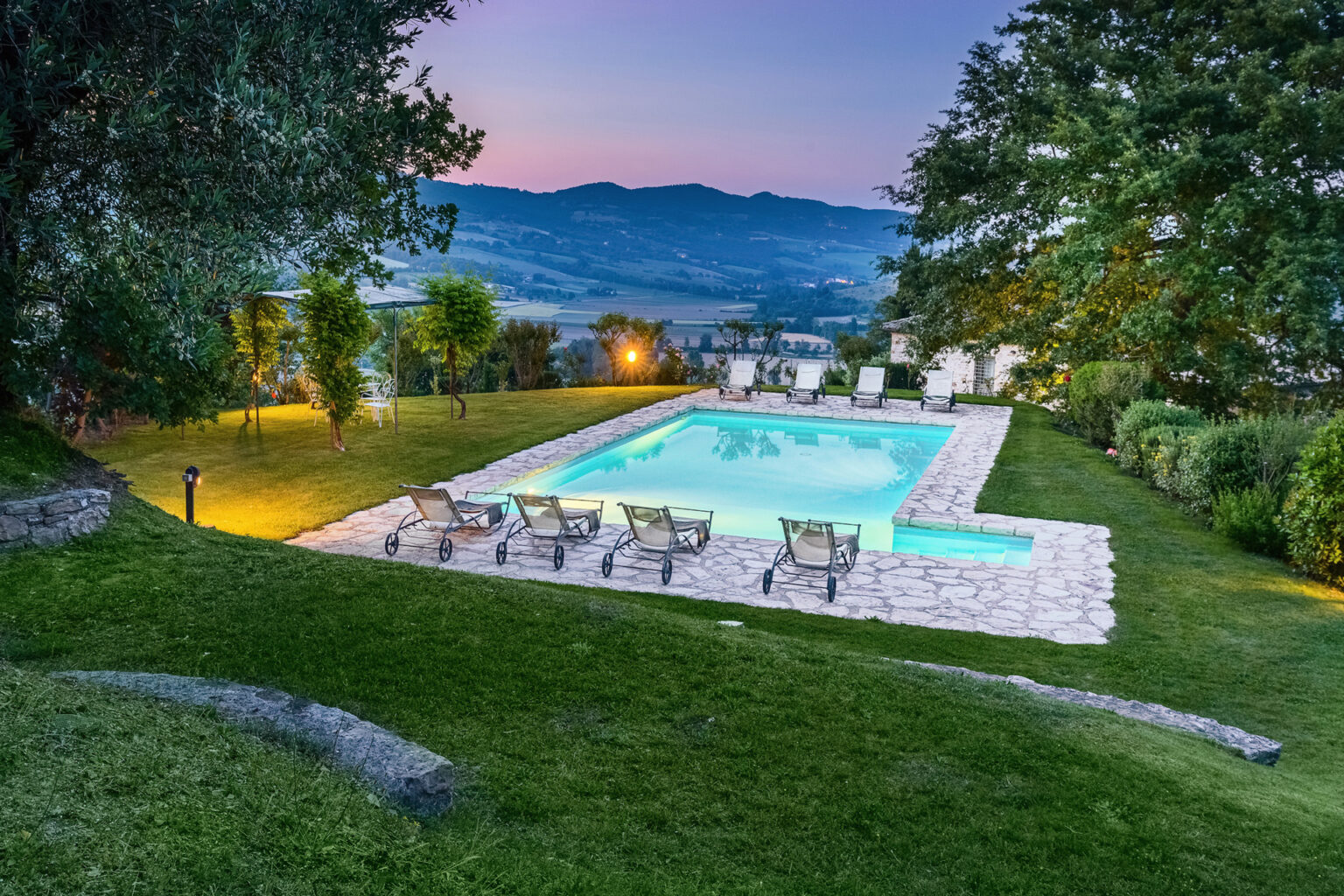 Scenic view of a serene outdoor pool at dusk, surrounded by lush greenery and lounge chairs, with rolling hills in the background. Perfect for relaxation and leisure. | Only on SopranoVillas