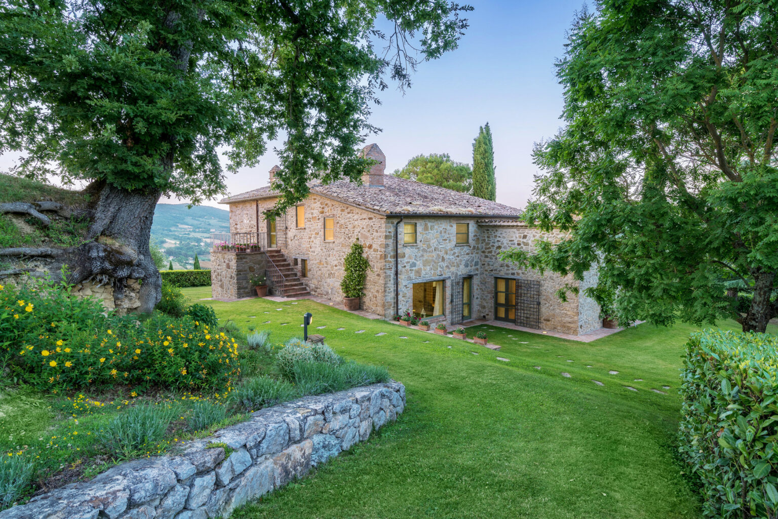 Charming stone villa surrounded by lush greenery and vibrant flowers, set against a picturesque hillside landscape. The two-story home features large windows, a tiled roof, and a spacious lawn, perfect for outdoor relaxation. | Only on SopranoVillas