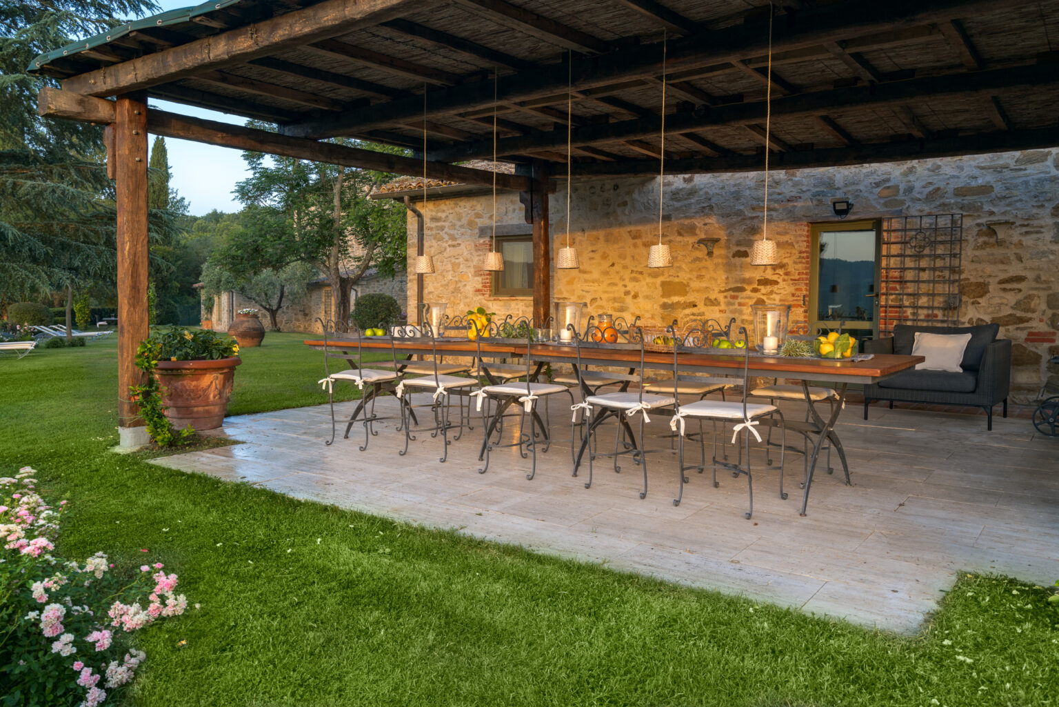 Outdoor dining area with a long wooden table and chairs, surrounded by lush green grass and blooming flowers, under a rustic covered patio. The setting is illuminated by soft lighting, creating a warm and inviting atmosphere for gatherings. | Only on SopranoVillas