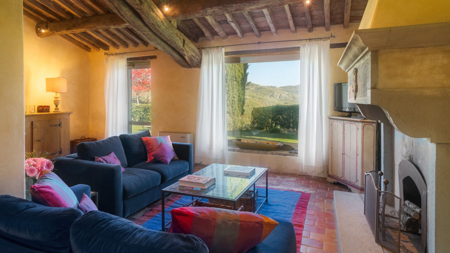Cozy living room with rustic decor, featuring a blue sofa with colorful pillows, a glass coffee table, and a view of greenery through large windows. Exposed wooden beams and warm lighting create an inviting atmosphere. | Only on SopranoVillas