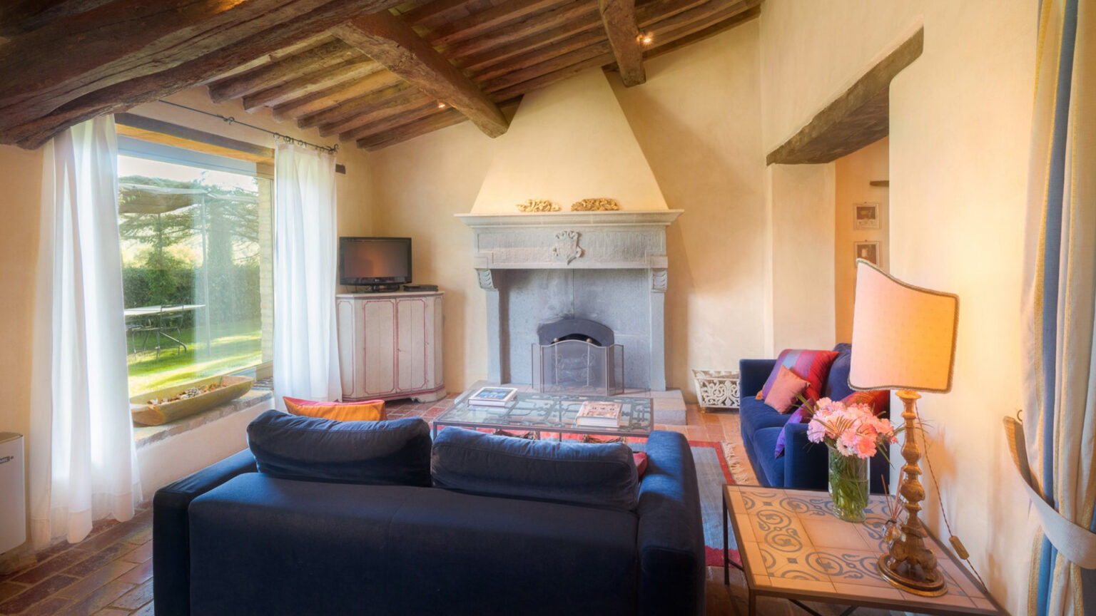 Cozy living room featuring a stone fireplace, comfortable blue sofas, a stylish lamp, and large windows offering natural light and a view of the outdoors. | Only on SopranoVillas