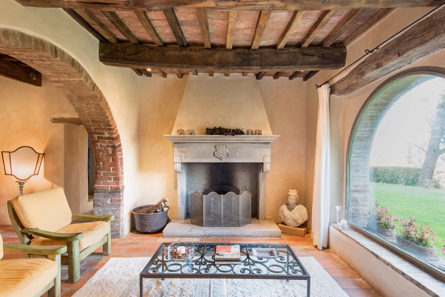 Cozy living room featuring a stone fireplace, wooden beam ceiling, and large window with garden view, complemented by a stylish coffee table and comfortable seating. | Only on SopranoVillas