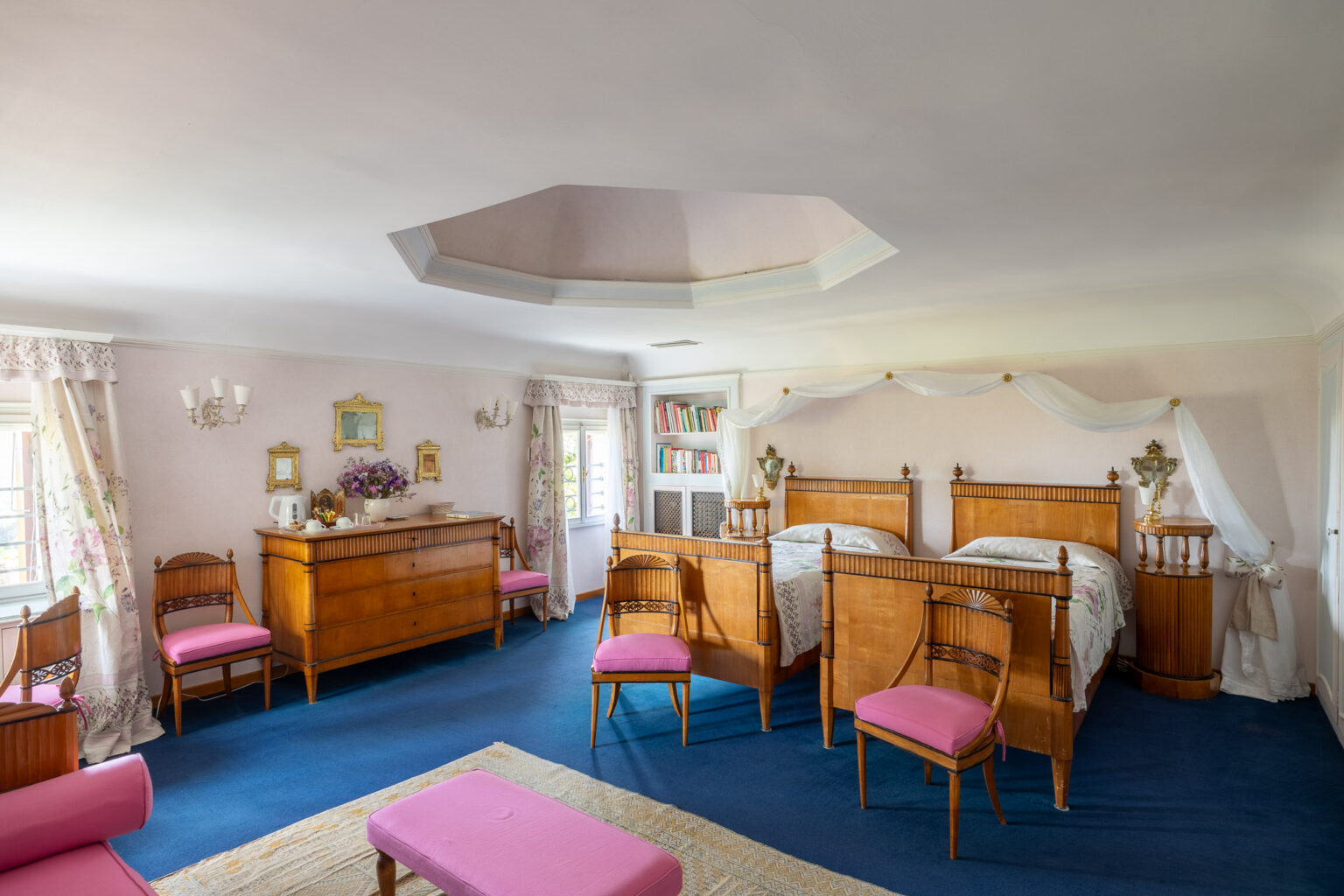Spacious and elegantly designed bedroom featuring two wooden beds with decorative headboards, pink upholstered chairs, a wooden dresser, and floral curtains. The room is adorned with vintage decor, including framed mirrors and a bookshelf, creating a warm and inviting atmosphere. | Only on SopranoVillas