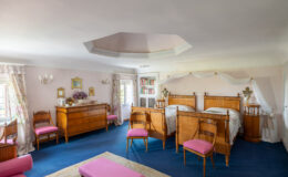 Spacious and elegantly designed bedroom featuring two wooden beds with decorative headboards, pink upholstered chairs, a wooden dresser, and floral curtains. The room is adorned with vintage decor, including framed mirrors and a bookshelf, creating a warm and inviting atmosphere.