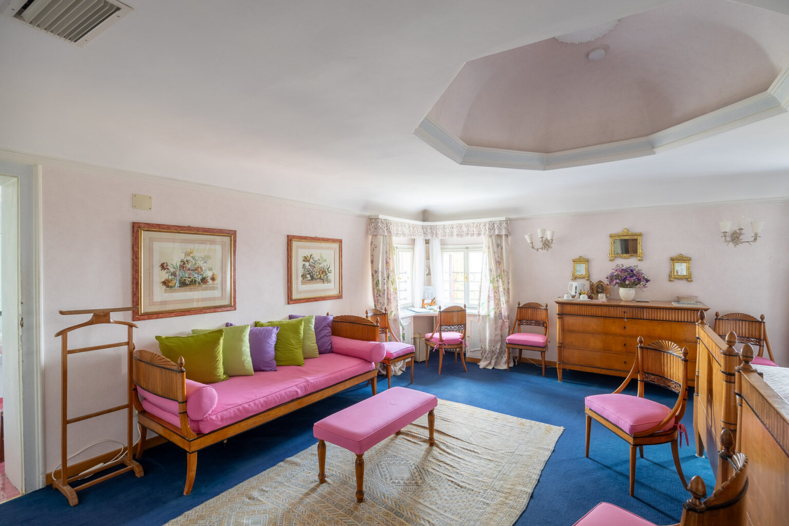 Bright and elegantly decorated living room featuring a pink and green color scheme, with a pink sofa adorned with colorful cushions, a matching bench, and wooden furniture. The room is illuminated by natural light from multiple windows, complemented by floral curtains and framed artwork on the walls. A cozy atmosphere is created by the plush carpet and decorative elements, making it an inviting space for relaxation. | Only on SopranoVillas