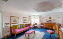 Bright and elegantly decorated living room featuring a pink and green color scheme, with a pink sofa adorned with colorful cushions, a matching bench, and wooden furniture. The room is illuminated by natural light from multiple windows, complemented by floral curtains and framed artwork on the walls. A cozy atmosphere is created by the plush carpet and decorative elements, making it an inviting space for relaxation.
