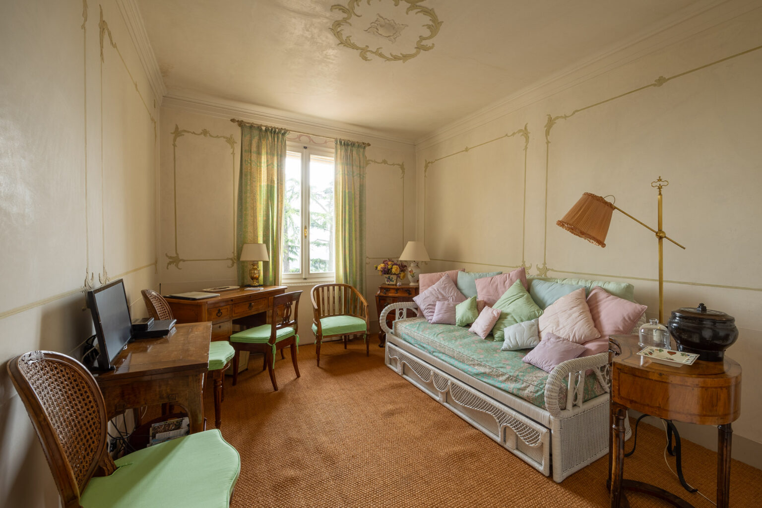 Cozy and elegantly decorated living room featuring a pastel-colored sofa adorned with various pillows, a wooden desk and chair, and a stylish floor lamp, all complemented by large windows with green curtains that allow natural light to brighten the space. | Only on SopranoVillas
