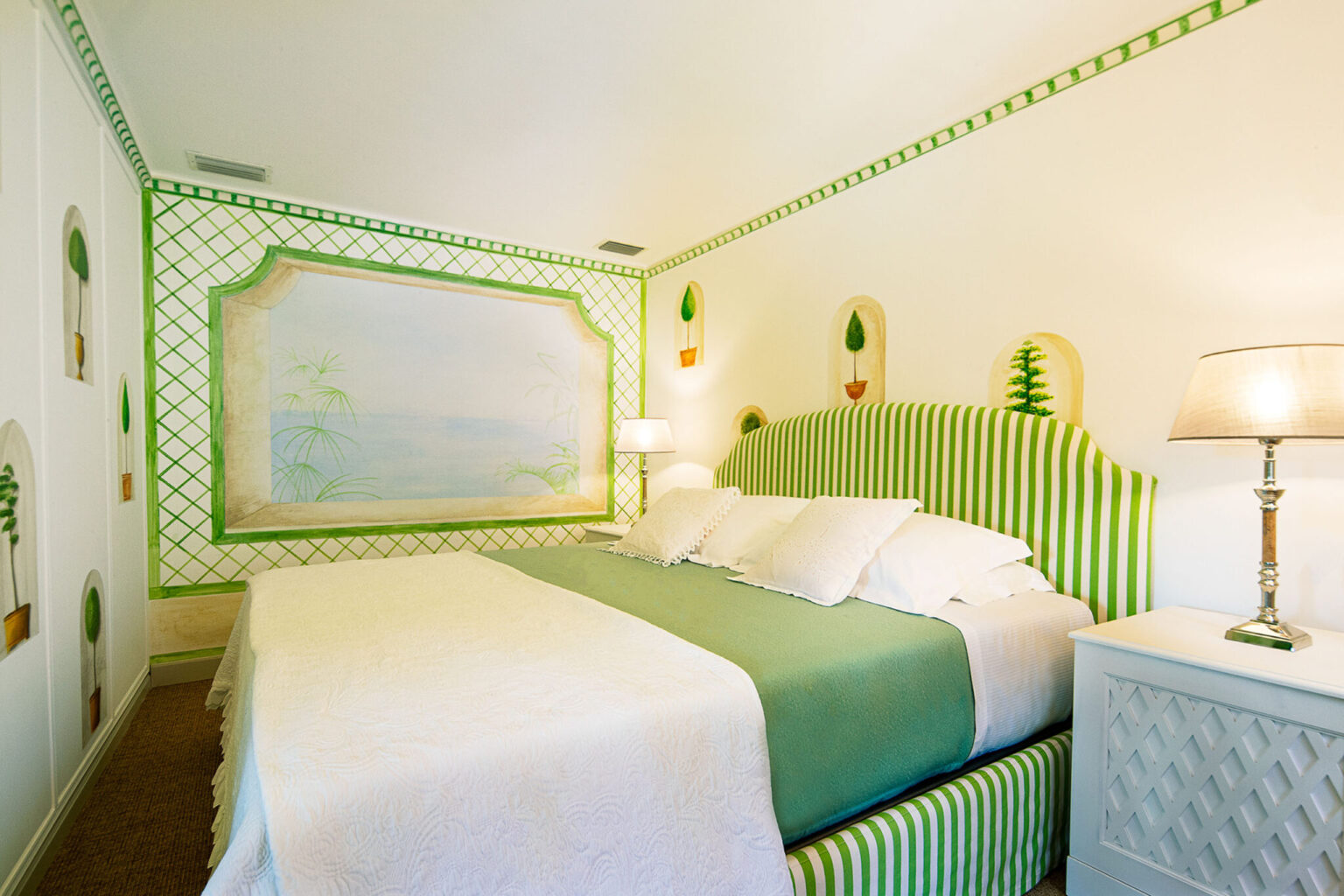 Cozy bedroom featuring a striped green and white headboard, a light green bedspread, and decorative pillows. The walls are adorned with a soft mural and potted plant designs, complemented by a modern lamp and a stylish nightstand. Ideal for a relaxing getaway. | Only on SopranoVillas