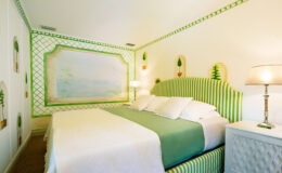Cozy bedroom featuring a striped green and white headboard, a light green bedspread, and decorative pillows. The walls are adorned with a soft mural and potted plant designs, complemented by a modern lamp and a stylish nightstand. Ideal for a relaxing getaway.
