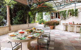 Outdoor dining area featuring a stone table set with various dishes, fruits, and beverages. Surrounded by lush greenery and decorative plants, the space is shaded by a canopy, creating a serene atmosphere ideal for meals.