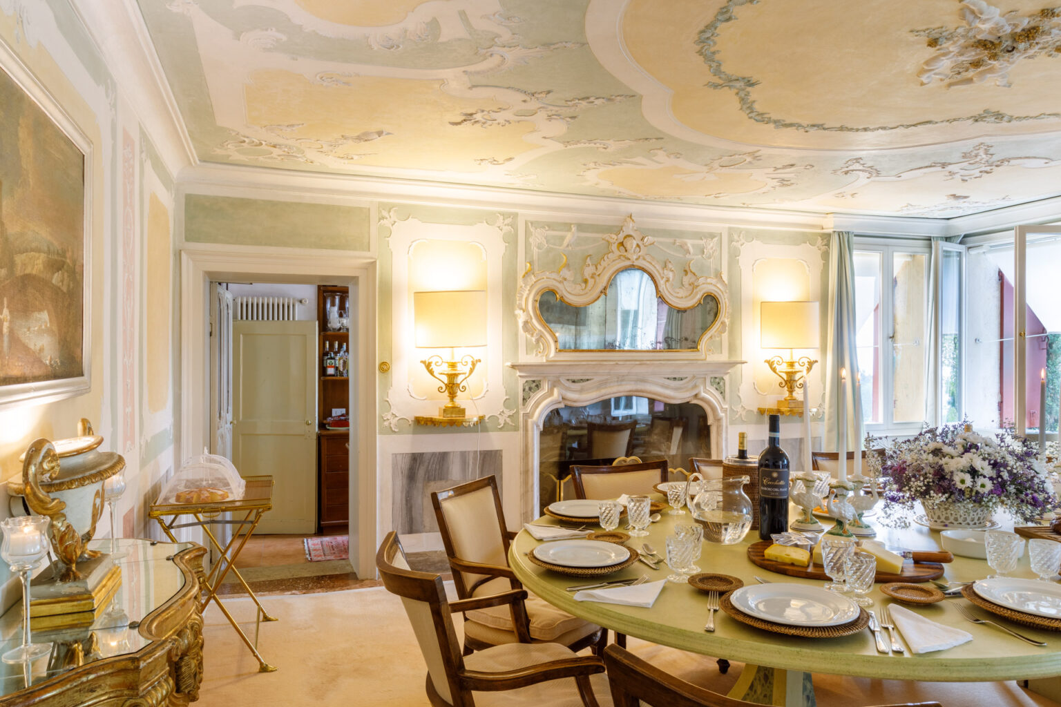 Elegant dining room featuring a round table set with fine china, crystal glassware, and a wine bottle. The decor includes ornate ceiling designs, a vintage mirror, and decorative wall accents, creating a sophisticated ambiance. Natural light streams in through large windows, enhancing the inviting atmosphere. | Only on SopranoVillas