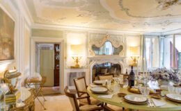 Elegant dining room featuring a round table set with fine china, crystal glassware, and a wine bottle. The decor includes ornate ceiling designs, a vintage mirror, and decorative wall accents, creating a sophisticated ambiance. Natural light streams in through large windows, enhancing the inviting atmosphere.