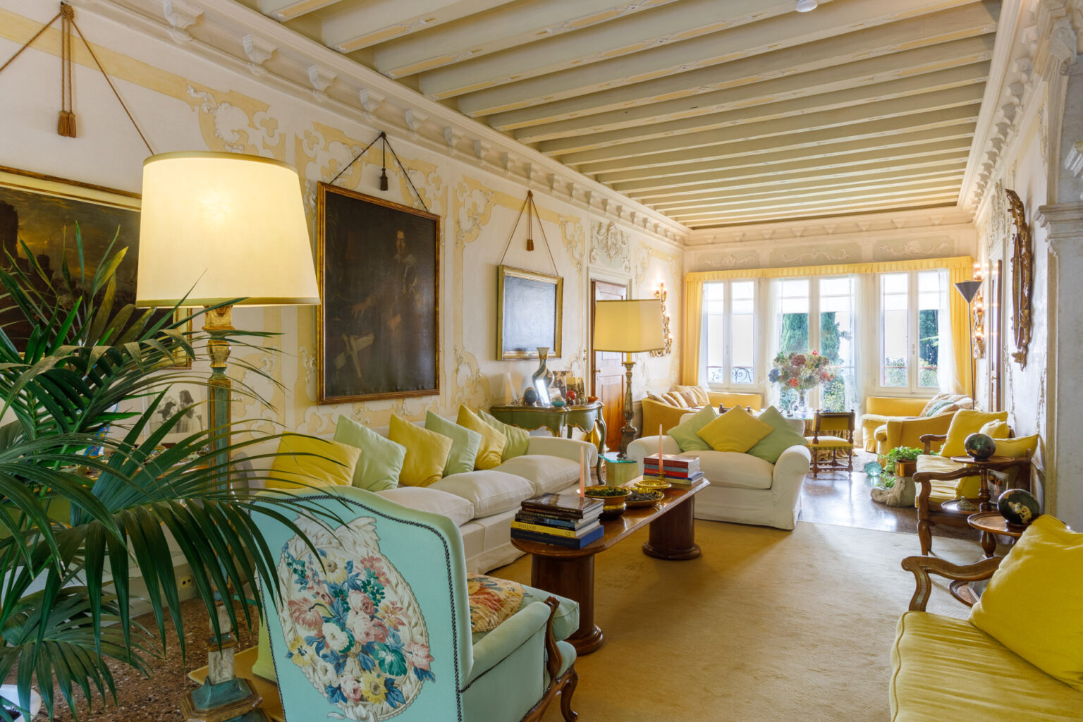 Bright and inviting living room featuring elegant decor, soft yellow accents, and vintage furniture. The space includes a comfortable white sofa adorned with pastel cushions, a decorative lamp, and art pieces on the walls. Large windows allow natural light to illuminate the room, enhancing the warm atmosphere. | Only on SopranoVillas