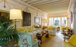 Bright and inviting living room featuring elegant decor, soft yellow accents, and vintage furniture. The space includes a comfortable white sofa adorned with pastel cushions, a decorative lamp, and art pieces on the walls. Large windows allow natural light to illuminate the room, enhancing the warm atmosphere.