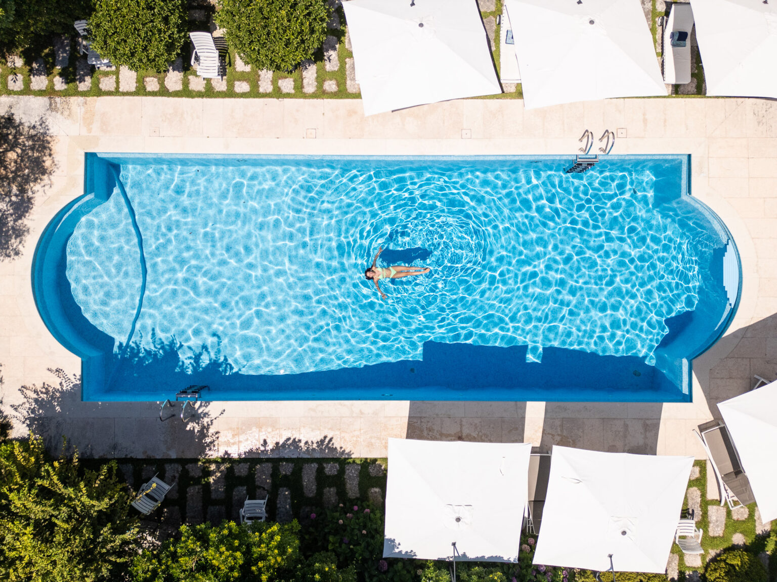 Aerial view of a serene swimming pool with a person floating on the water, surrounded by lounge chairs and shaded umbrellas, set in a lush garden environment. | Only on SopranoVillas