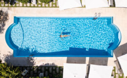 Aerial view of a serene swimming pool with a person floating on the water, surrounded by lounge chairs and shaded umbrellas, set in a lush garden environment.