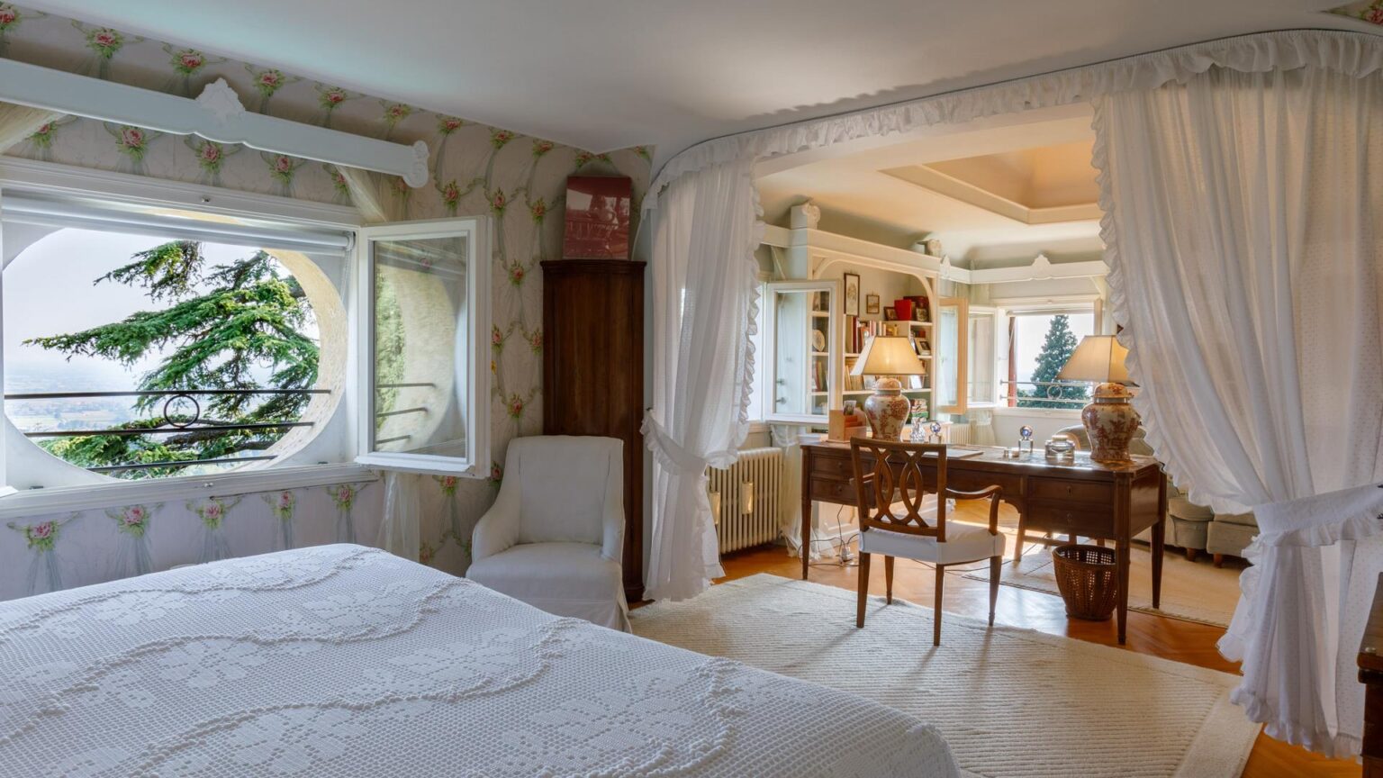 Cozy bedroom with a white bedspread, featuring a large circular window showcasing a scenic view of greenery. The room includes a comfortable chair, a wooden desk with elegant lamps, and decorative elements like floral wallpaper and a plush area rug, creating a warm and inviting atmosphere. | Only on SopranoVillas