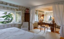 Cozy bedroom with a white bedspread, featuring a large circular window showcasing a scenic view of greenery. The room includes a comfortable chair, a wooden desk with elegant lamps, and decorative elements like floral wallpaper and a plush area rug, creating a warm and inviting atmosphere.