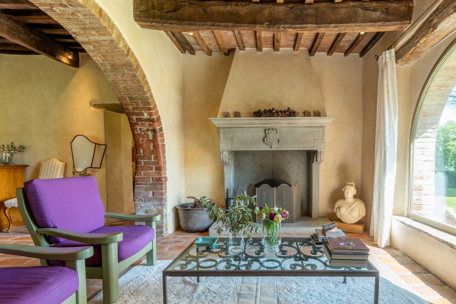 Cozy living room with rustic wooden beams, a stone fireplace, purple chairs, and a glass coffee table adorned with flowers, featuring large windows that allow natural light to flood the space. | Only on SopranoVillas