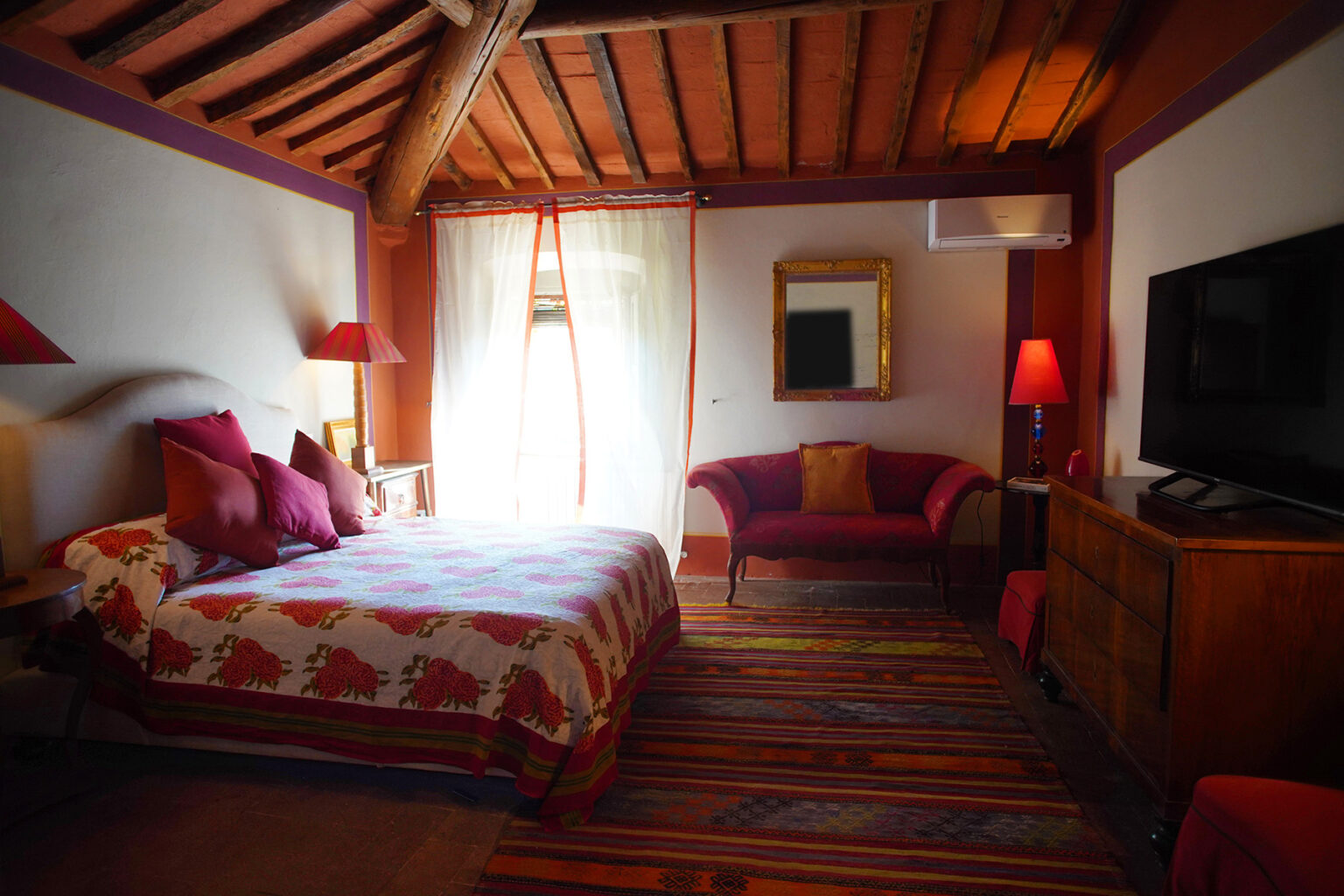 Cozy hotel room featuring a queen-sized bed with decorative pillows, a vibrant area rug, a stylish red sofa, and a flat-screen TV, all set against a backdrop of wooden beams and natural light. | Only on SopranoVillas