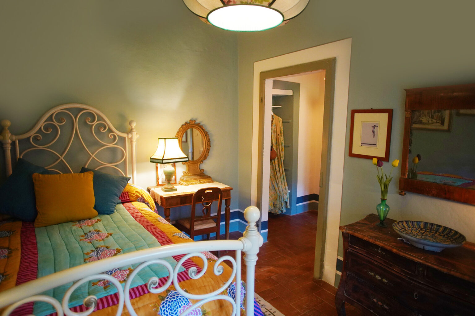 Cozy bedroom featuring a decorative metal bed with colorful bedding, a bedside lamp, and a wooden table with a mirror. The room has a warm ambiance with soft lighting and a view of an adjoining room. | Only on SopranoVillas