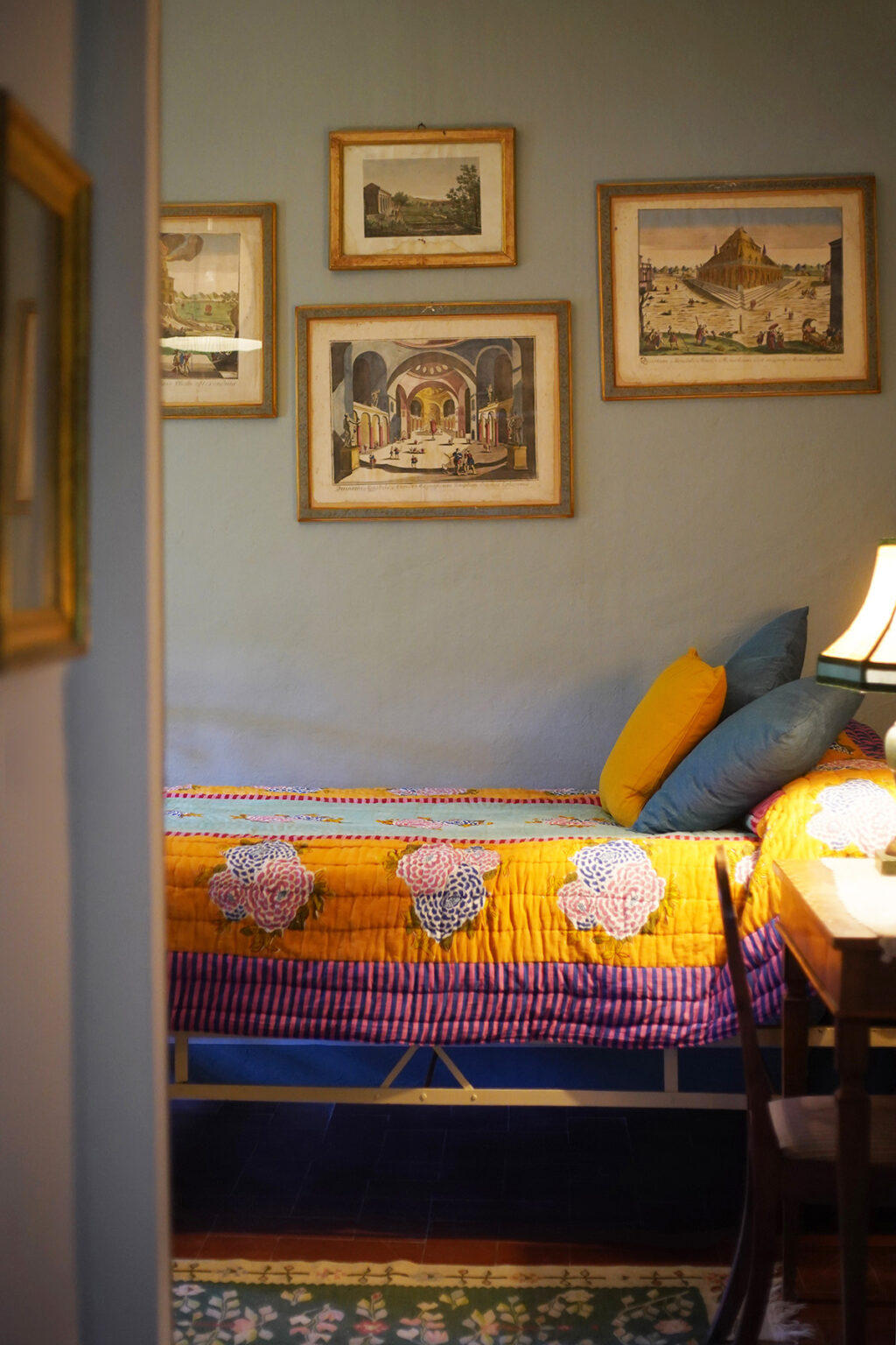 Cozy bedroom interior featuring a colorful quilt with floral patterns, decorative pillows in blue and yellow, and framed artwork on the wall, creating a warm and inviting atmosphere. | Only on SopranoVillas