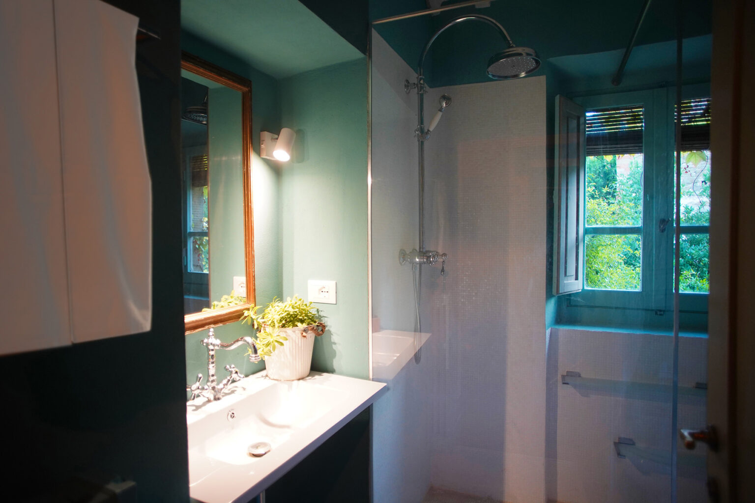 Modern bathroom interior featuring a shower, sink with vanity, and a window with greenery view. The walls are painted in a soothing teal color, complemented by stylish fixtures and natural light. | Only on SopranoVillas