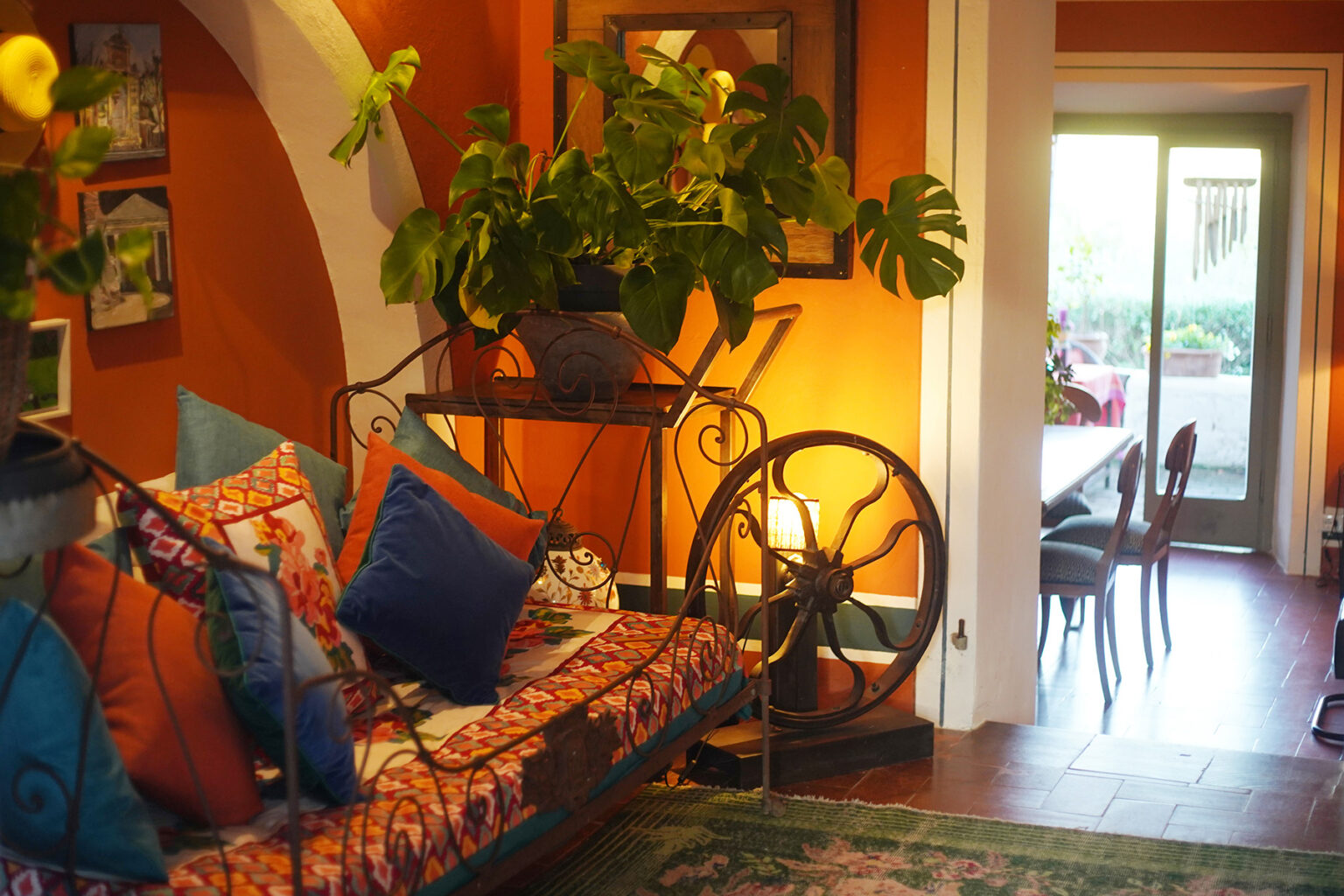 Cozy living room featuring vibrant orange walls, a decorative couch with colorful cushions, and a large potted plant. The space is illuminated by warm lighting and leads to a dining area with wooden chairs. | Only on SopranoVillas