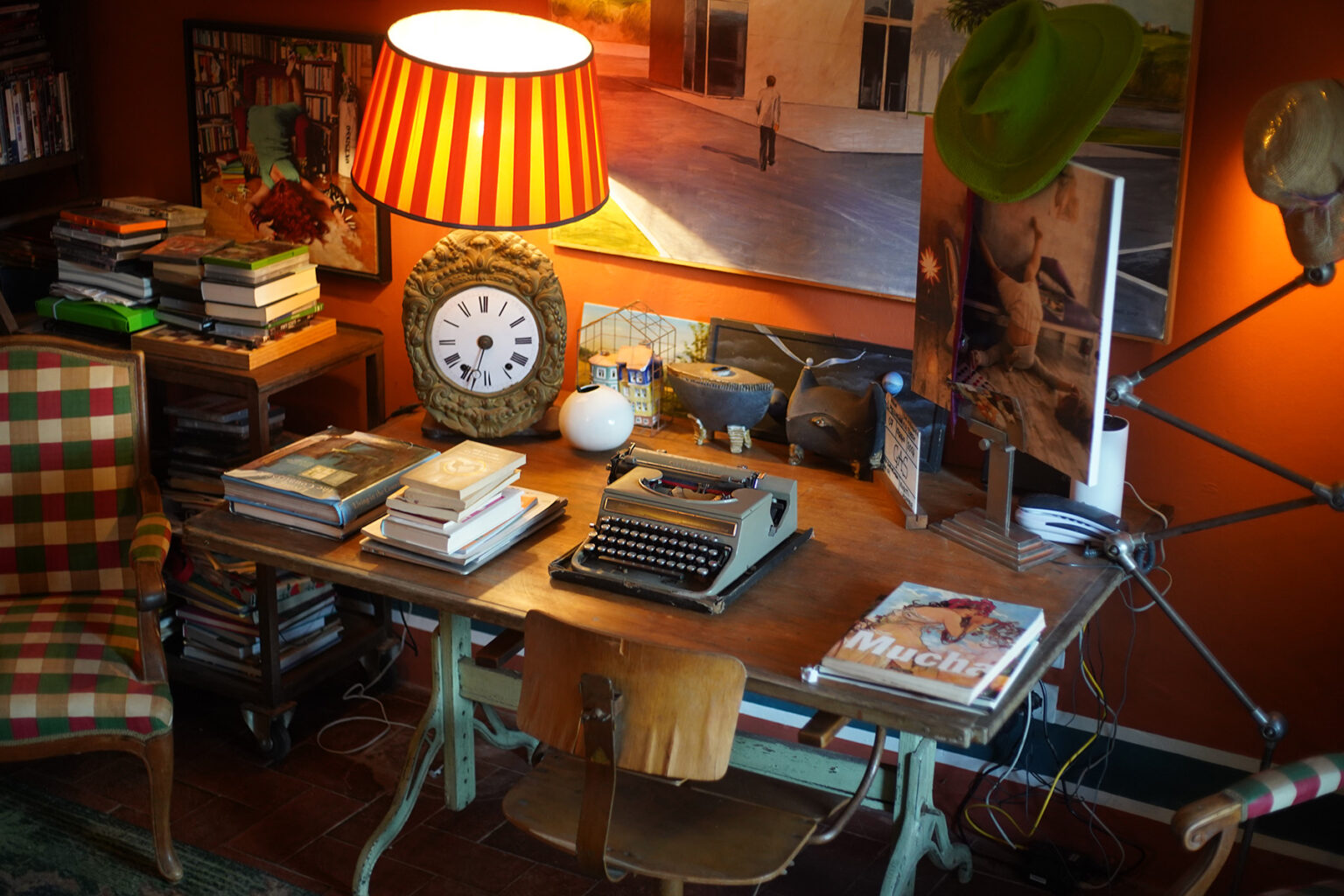 Cozy vintage workspace featuring a wooden desk with a typewriter, stacks of books, a decorative clock, and a striped lamp, surrounded by warm orange walls and artistic decor. | Only on SopranoVillas