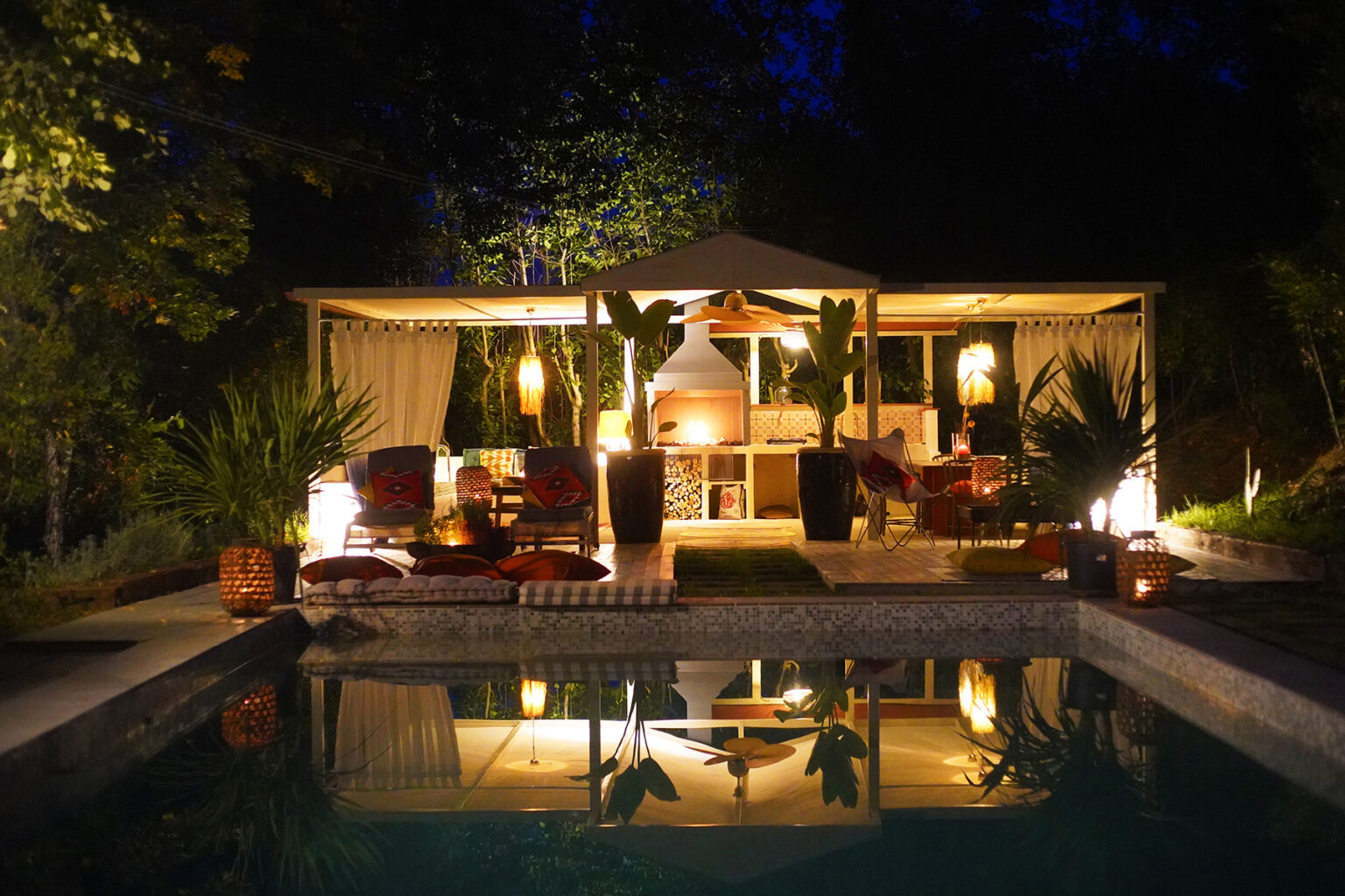 Elegant outdoor living space at night featuring a pool, cozy seating area with plush chairs, and soft lighting surrounded by lush greenery. | Only on SopranoVillas