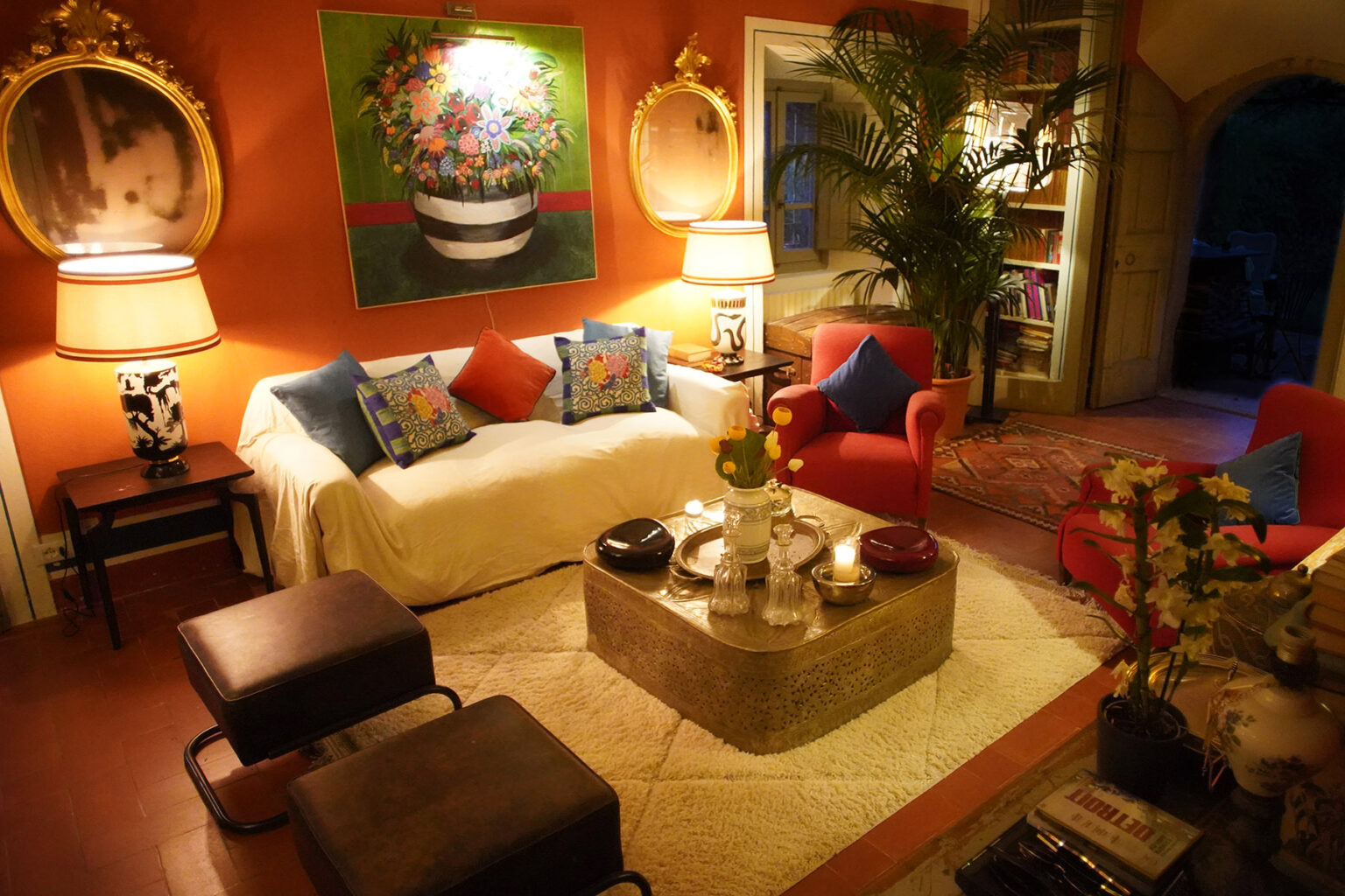 Cozy living room with vibrant orange walls, featuring a white sofa adorned with colorful pillows, red armchairs, a decorative coffee table, and artistic wall decor. The space is illuminated with warm lighting, creating an inviting atmosphere. | Only on SopranoVillas