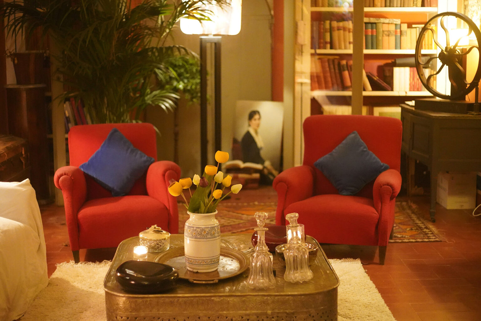 Cozy living room setup featuring two red armchairs with blue cushions, a coffee table adorned with a vase of yellow tulips, and decorative glass decanters. A warm ambiance is created by a standing lamp, with bookshelves and a framed portrait in the background. | Only on SopranoVillas