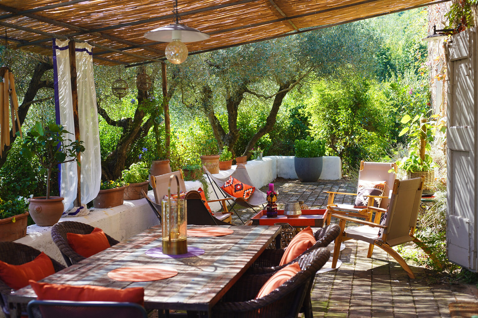 Cozy outdoor patio with a wooden table, comfortable seating, and lush greenery, featuring potted plants and a shaded area under a pergola, perfect for relaxation and dining. | Only on SopranoVillas