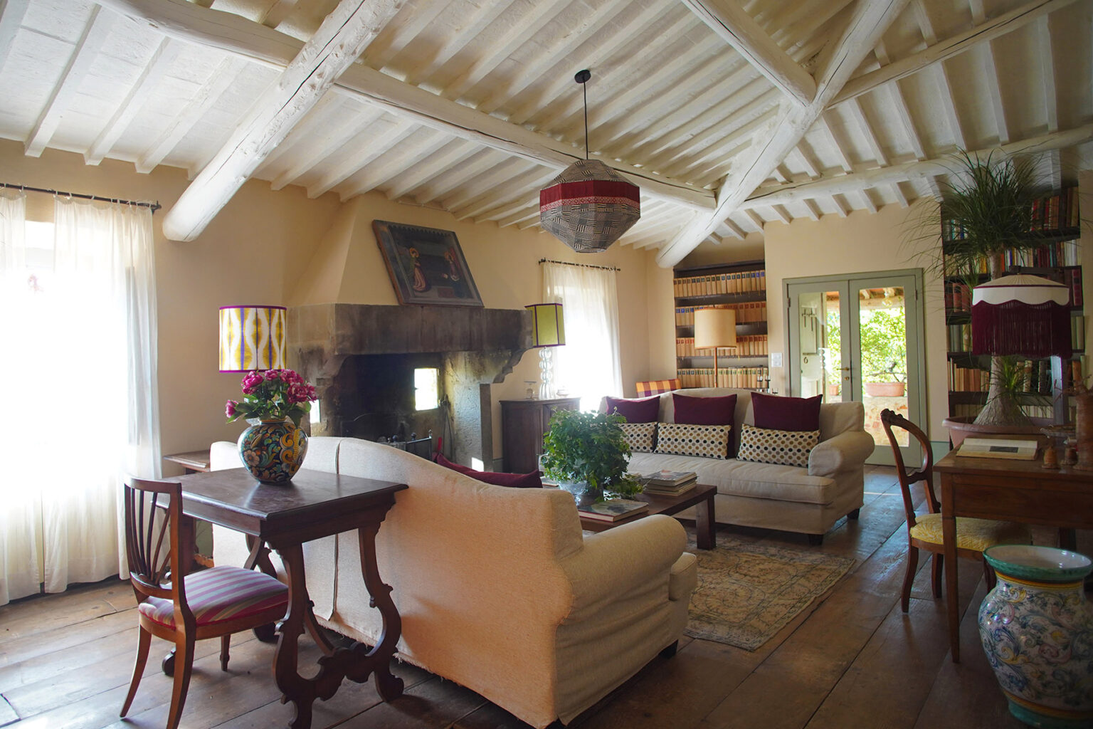 Bright and inviting living room featuring white sofas, a wooden coffee table, and a cozy fireplace. The room has a rustic design with exposed wooden beams and large windows allowing natural light. Decorated with plants and bookshelves, it embodies a warm and welcoming atmosphere. | Only on SopranoVillas