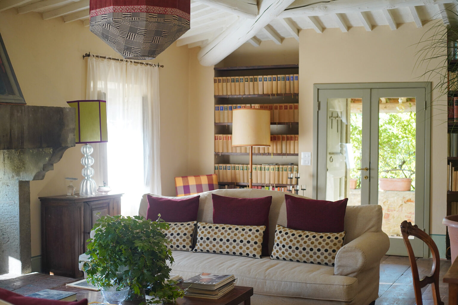 Cozy living room featuring a light-colored sofa with patterned cushions, a decorative lamp, a wooden side table, and a bookshelf filled with books. Natural light streams through sheer curtains, highlighting the rustic charm of the space with a stone fireplace and glass doors leading to an outdoor area. | Only on SopranoVillas