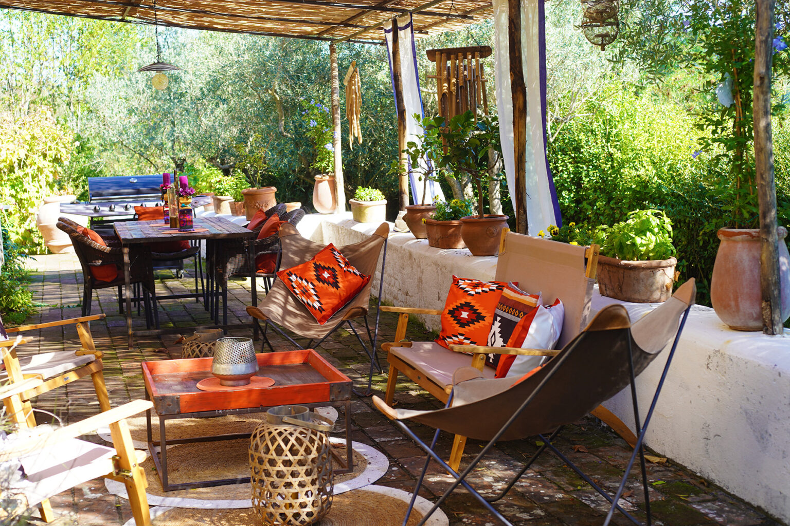 Cozy outdoor seating area featuring stylish chairs with vibrant cushions, a rustic wooden dining table, and decorative pots surrounded by lush greenery, perfect for entertaining or relaxing in nature. | Only on SopranoVillas