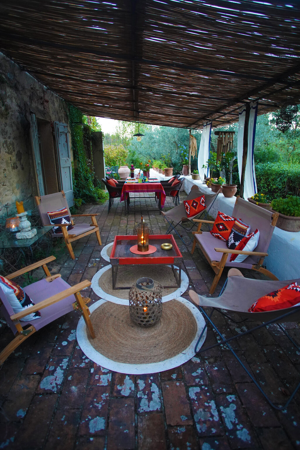 Cozy outdoor living space featuring wooden chairs with colorful cushions, a small table with a candle centerpiece, and a dining area set with a tablecloth and flowers, surrounded by greenery and rustic stone walls. | Only on SopranoVillas