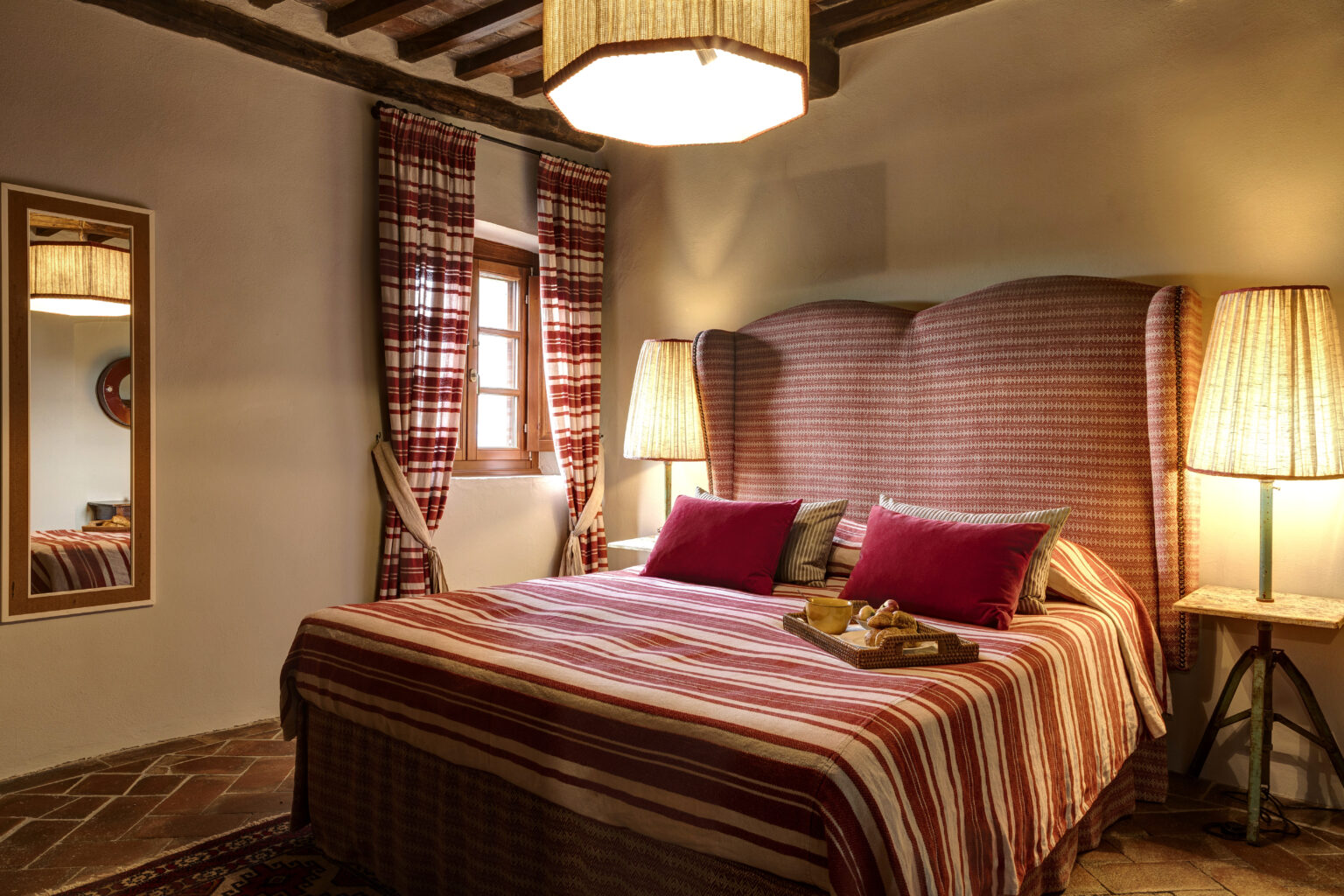 Cozy bedroom featuring a striped bedspread, plush red pillows, and a stylish high-backed headboard. The room is illuminated by a unique pendant light and two bedside lamps, complemented by checkered curtains and a mirror reflecting a warm, inviting atmosphere. A tray with pastries adds a touch of elegance to the space. | Only on SopranoVillas