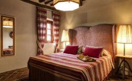 Cozy bedroom featuring a striped bedspread, plush red pillows, and a stylish high-backed headboard. The room is illuminated by a unique pendant light and two bedside lamps, complemented by checkered curtains and a mirror reflecting a warm, inviting atmosphere. A tray with pastries adds a touch of elegance to the space.