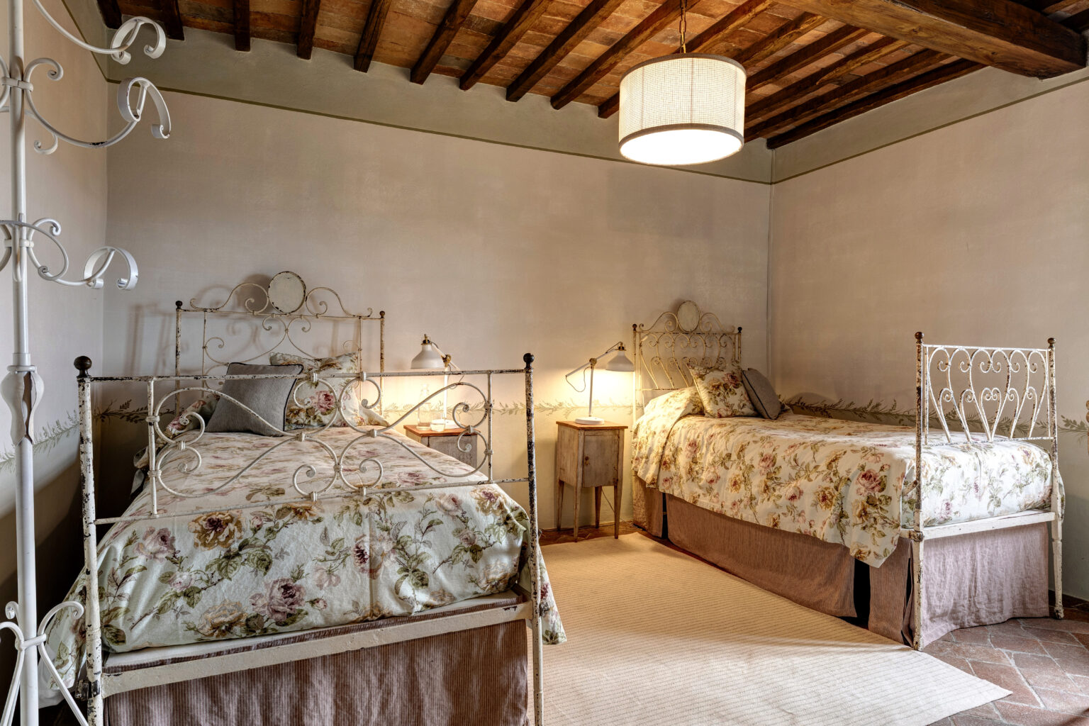 Cozy guest bedroom featuring two vintage iron beds with floral-patterned bedding, a warm atmosphere created by bedside lamps, and rustic wooden ceiling beams. The room is adorned with soft textures and a neutral color palette, ideal for a relaxing retreat. | Only on SopranoVillas