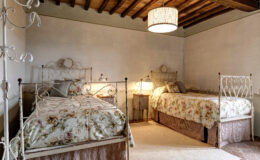 Cozy guest bedroom featuring two vintage iron beds with floral-patterned bedding, a warm atmosphere created by bedside lamps, and rustic wooden ceiling beams. The room is adorned with soft textures and a neutral color palette, ideal for a relaxing retreat.