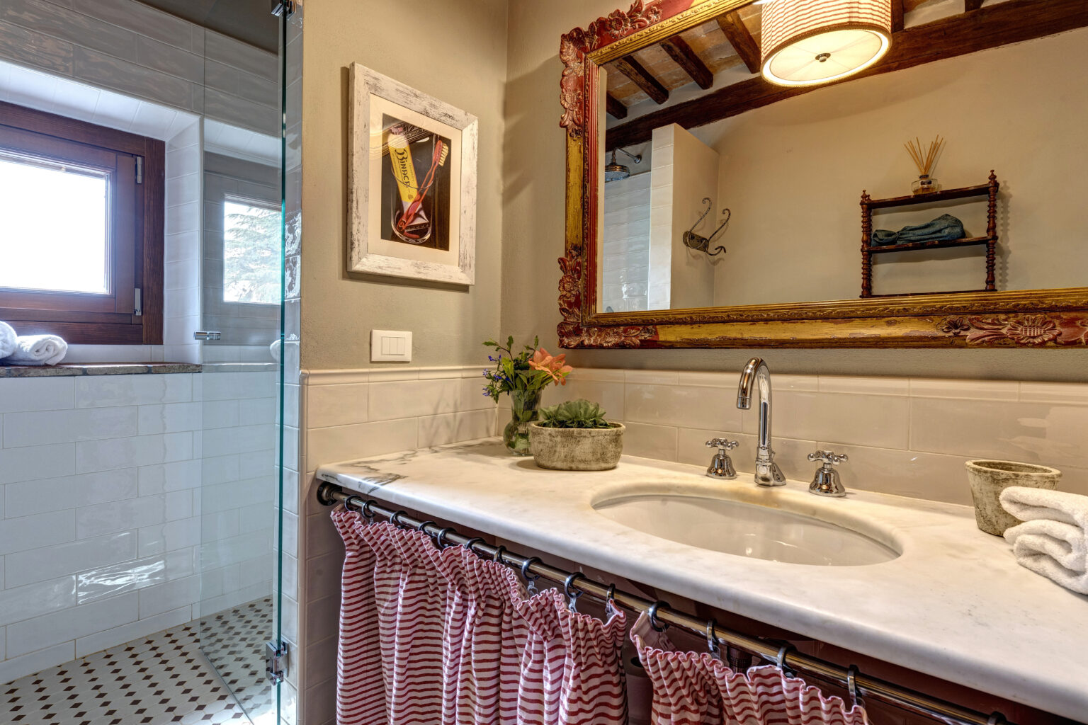 Modern bathroom interior featuring a marble sink with chrome faucets, decorative mirror, and a vibrant flower arrangement. The space includes a glass shower enclosure, wooden window, and striped curtain, creating a stylish and inviting atmosphere. | Only on SopranoVillas