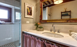 Modern bathroom interior featuring a marble sink with chrome faucets, decorative mirror, and a vibrant flower arrangement. The space includes a glass shower enclosure, wooden window, and striped curtain, creating a stylish and inviting atmosphere.